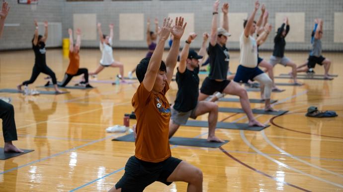 A group of students does yoga in a gym. They are in warrior pose. 